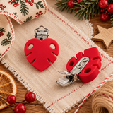 Deux pinces d’attache-tétine en silicone rouge avec attaches métalliques argentées, présentées sous différents angles sur un flatlay de Noël aux tons naturels, avec tissu festif, branches de sapin et étoile en bois.